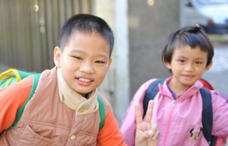 Zwei taubstumme asiatische Schulkinder mit Rucksäcken lächeln in die Kamera.